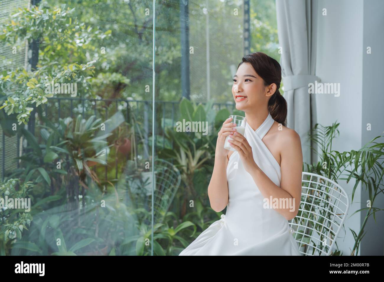 Una señora asiática bebiendo un vaso de leche mientras mira hacia fuera ventana con espacio de copia Foto de stock