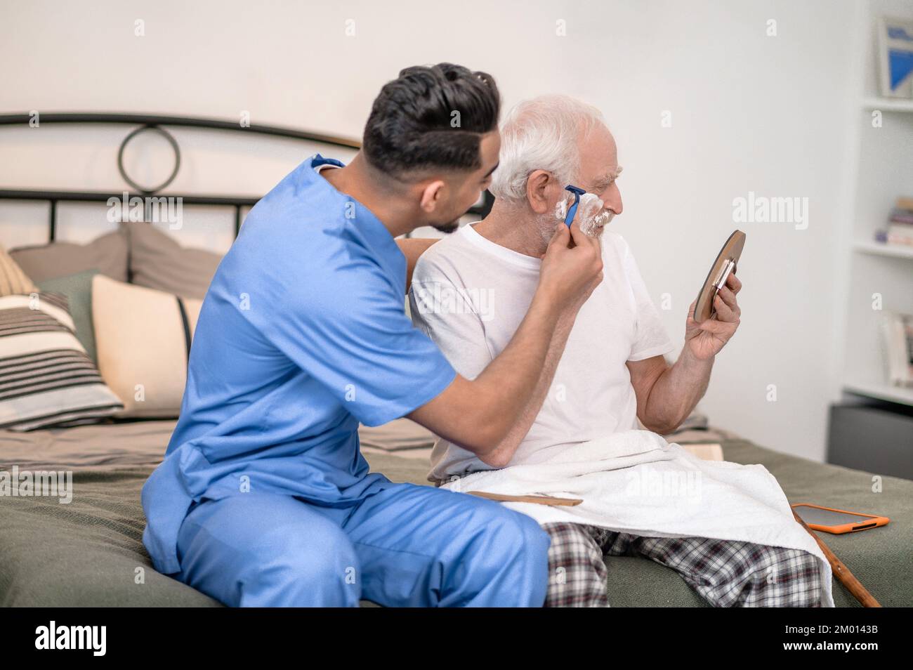 Hombre anciano mirándose a sí mismo en el espejo mientras su cuidador tira de la cuchilla a lo largo de sus bigotes. Foto de stock