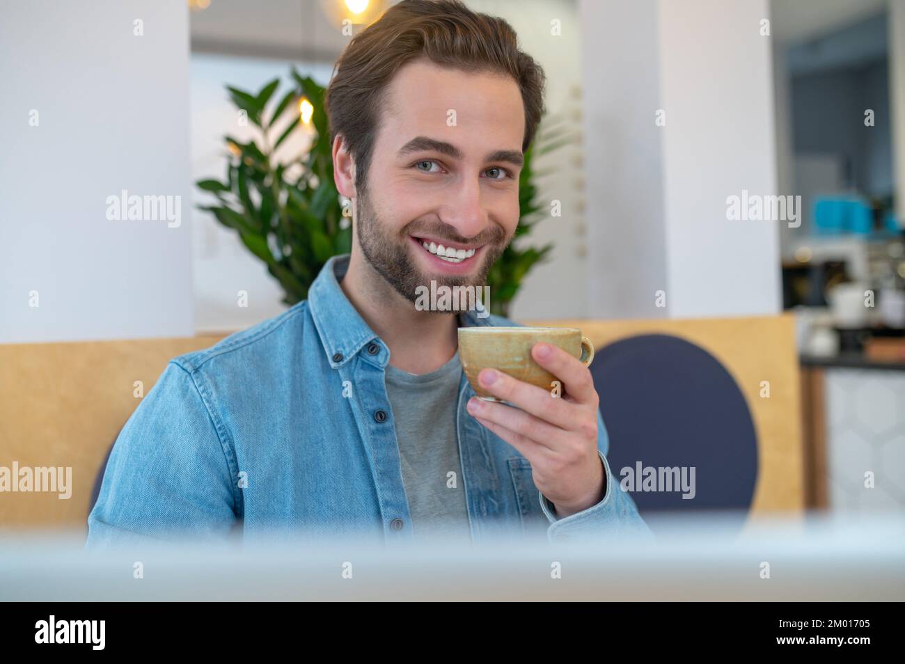 Todo excelente. Un hombre agradable y atractivo con una sonrisa tootea sentado sosteniendo la taza con confianza mirando la cámara en el café. Foto de stock