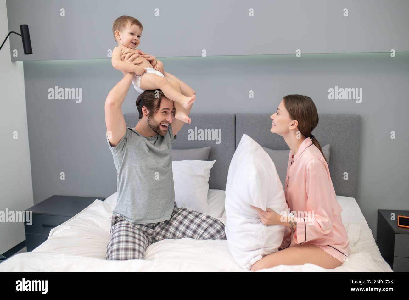 Humor divertido. Mujer sonriente en pijama con almohada sentada enfrente  mirando al hombre sosteniendo al bebé sobre la cabeza en la cama en el  dormitorio Fotografía de stock - Alamy