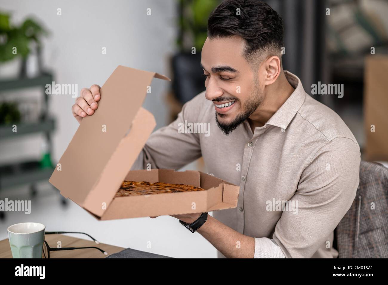 Disfrute. Feliz joven hombre barbudo con los ojos cerrados sosteniendo la caja de pizza abierta mientras se sienta en la mesa adentro. Foto de stock