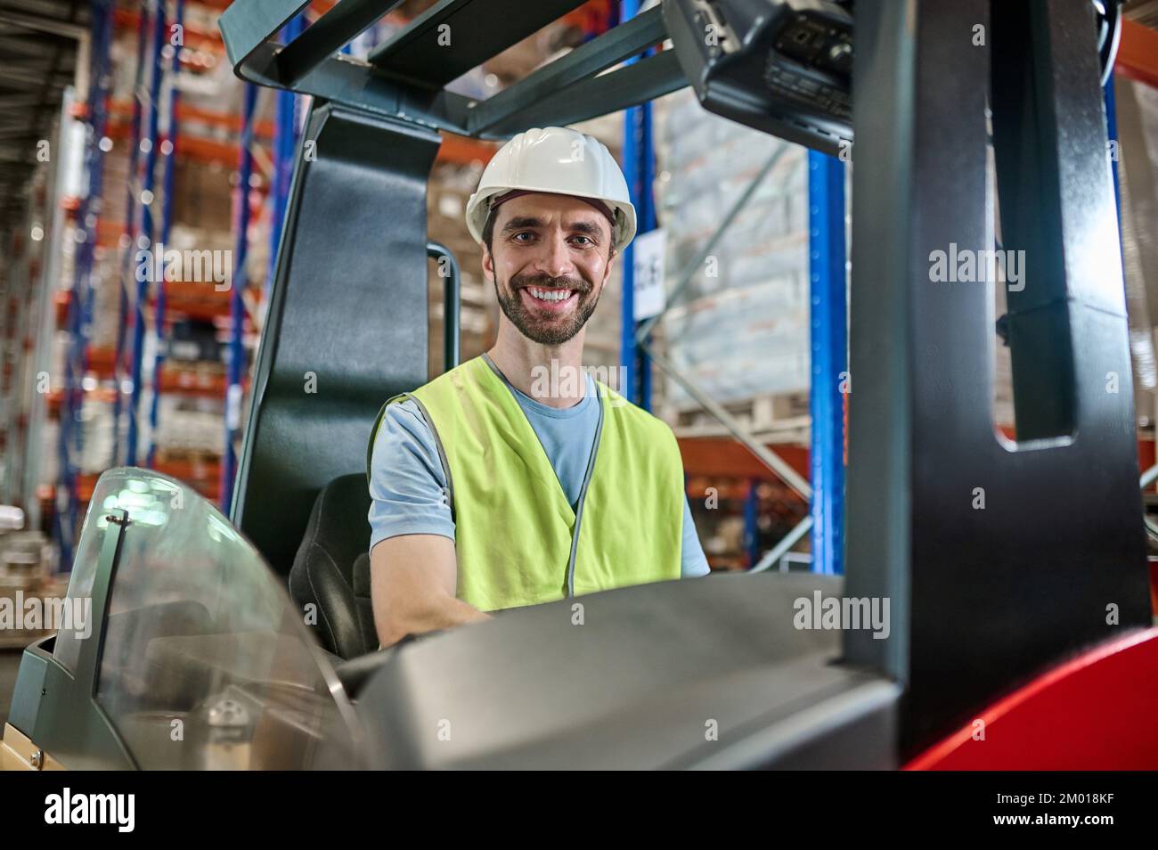 Sonriendo alegre trabajador de almacén en un casco y chaleco reflectante sentado en la carretilla elevadora eléctrica. Foto de stock
