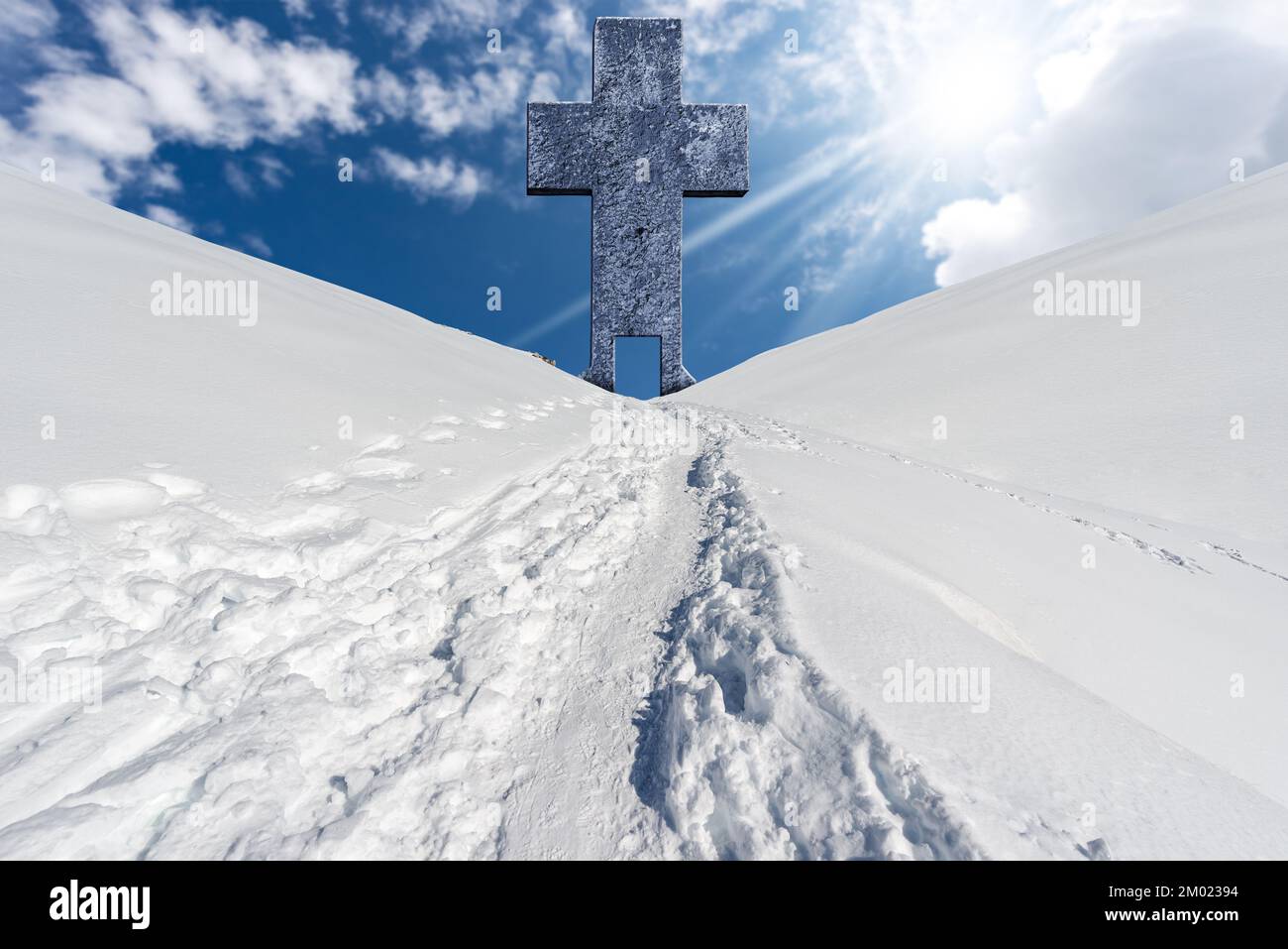 Gran cruz religiosa de piedra con una puerta y un sendero con huellas en la nieve en un paisaje de montaña contra un cielo azul con nubes y rayos de sol. Foto de stock