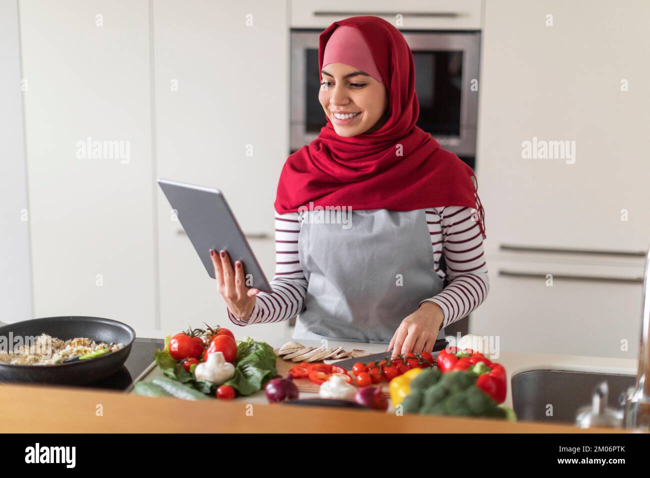 Sonriente dama musulmana cocinando en casa, utilizando la aplicación de cocina Foto de stock