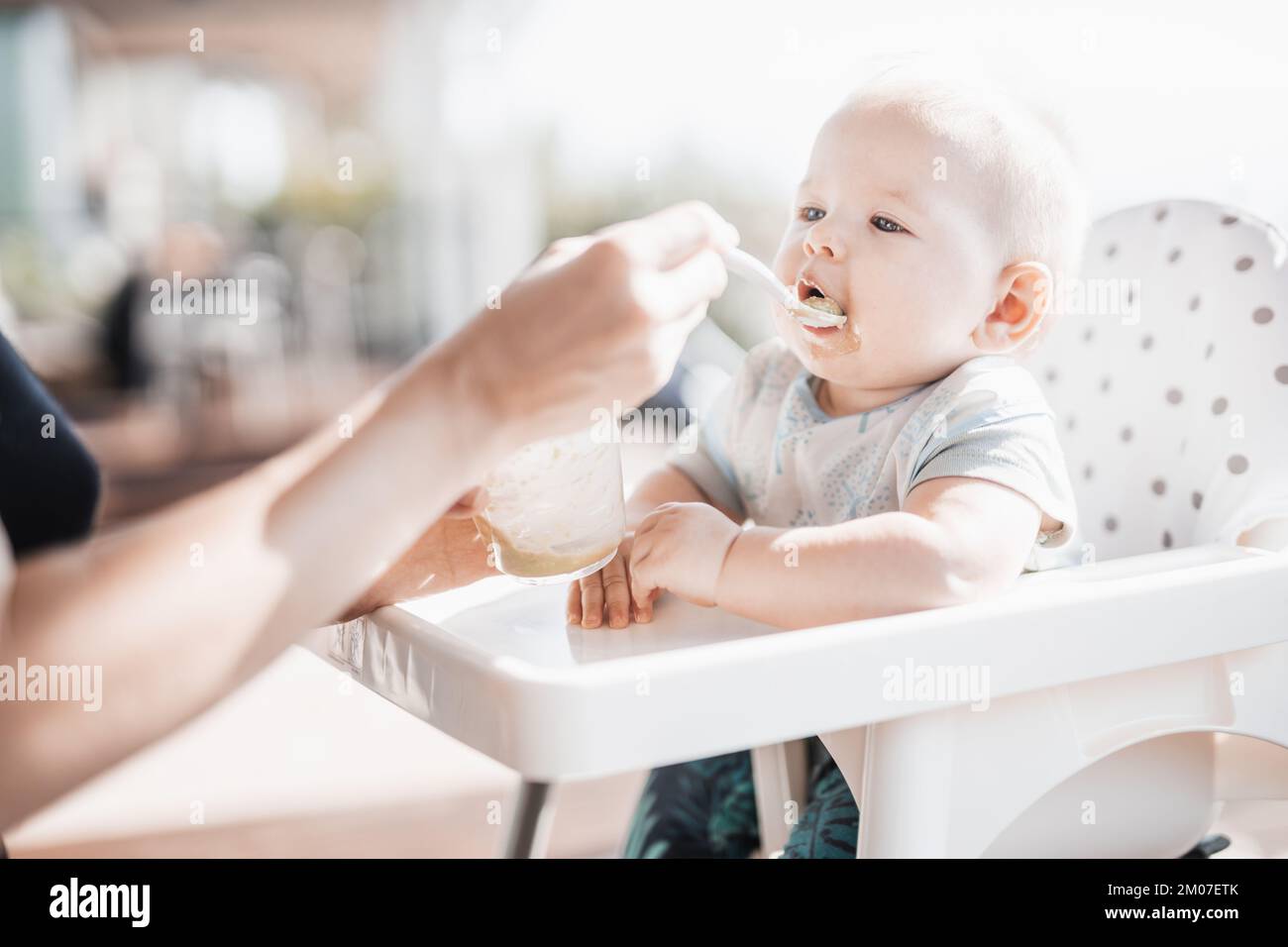 Madre cuchara alimentar a su bebé niño en silla de bebé con puré de frutas en un porche en vacaciones de verano. Concepto de introducción de alimentos sólidos para bebés Foto de stock