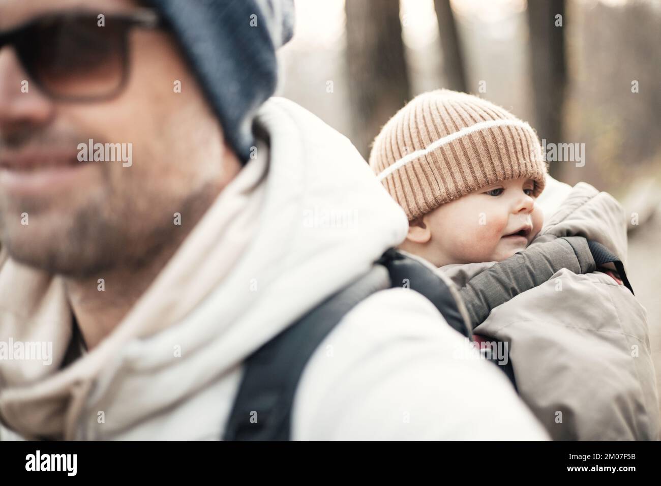 Padre deportivo llevando a su hijo pequeño con traje de mono de invierno y gorra en mochila de senderismo en el bosque de otoño Foto de stock