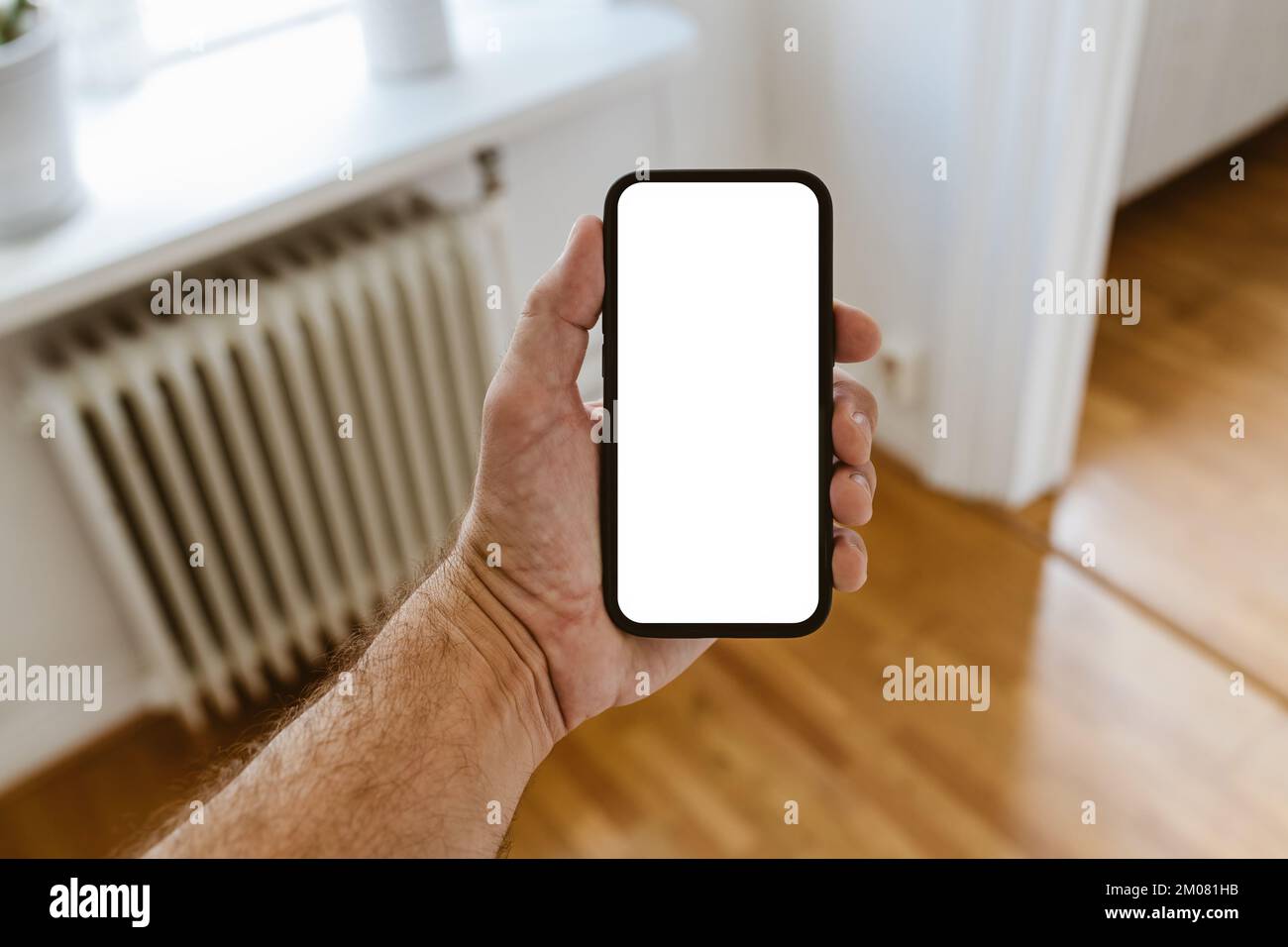 Mano masculina con mockup de teléfono móvil, hombre sosteniendo smartphone con pantalla de mockup en blanco en el salón de casa. Enfoque selectivo. Foto de stock