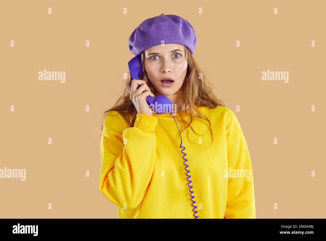 Aturdida mujer perpleja con sombrero púrpura y sudadera amarilla llamando al teléfono sobre fondo beige. Foto de stock