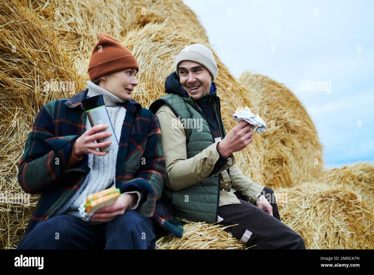 Dos trabajadores jóvenes felices de granja en ropa de trabajo sentados junto a la pila de heno delante de la cámara, hablando y tomando sándwiches y té para el almuerzo Foto de stock