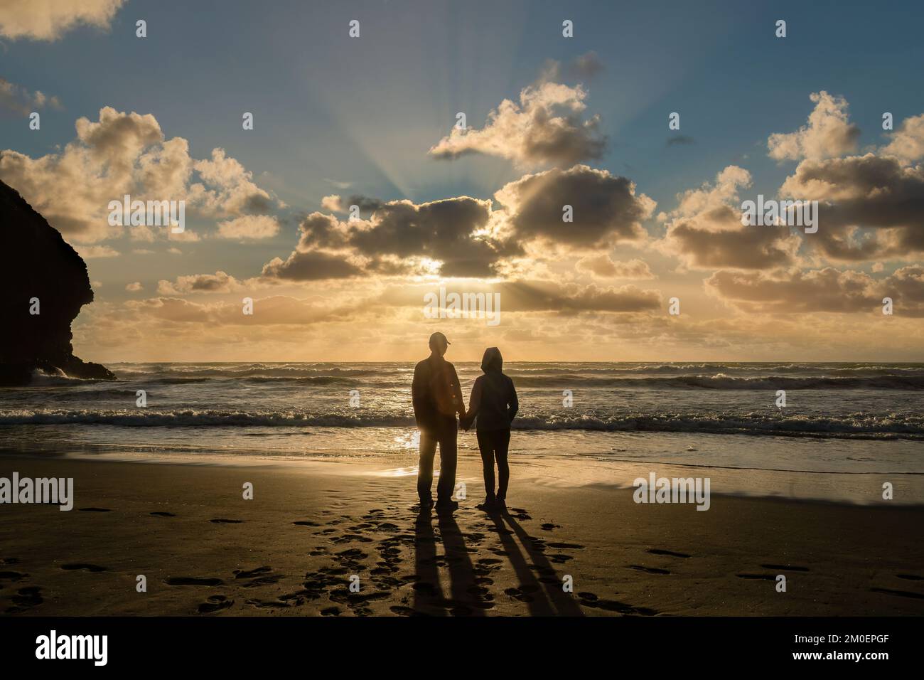 Una pareja viendo la puesta de sol en Piha Beach, rayos de sol brillando a  través de las nubes. Waitakere, Auckland Fotografía de stock - Alamy