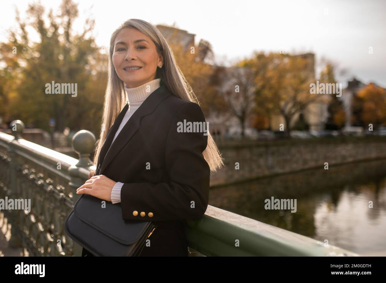 Mujer de mediana edad con un abrigo negro parado en el puente del río Foto de stock