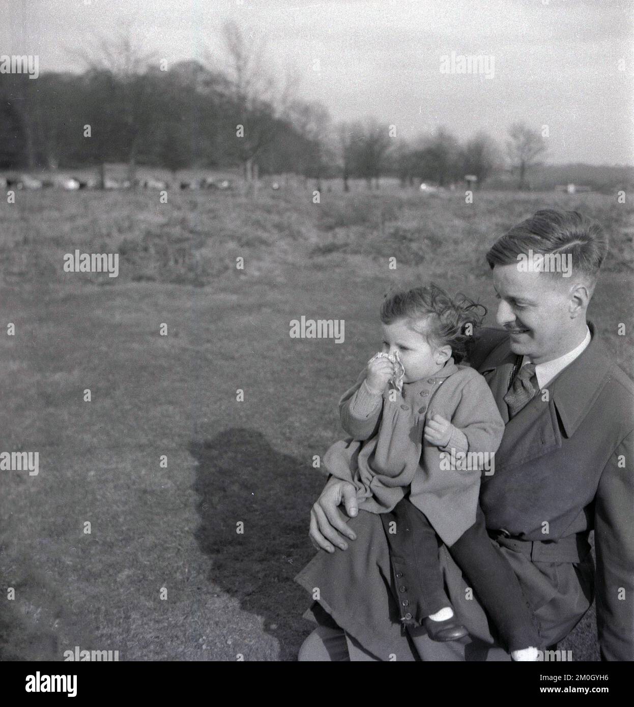 1950s, histórico, fuera en páramos abiertos, un padre que llevaba un impermeable con su hija pequeña descansando sobre su rodilla, Inglaterra, Reino Unido. Foto de stock