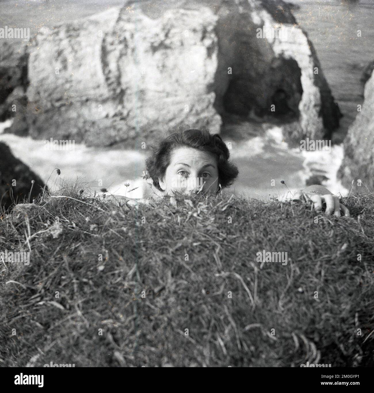 1950s, histórico, en la costa, en lo alto del océano, una dama ocultando la mitad de su rostro bajo el suelo, fingiendo tal vez que se está aferrando a la cara del acantilado o voladizo para sorprender a un amigo, Inglaterra, Reino Unido. Foto de stock