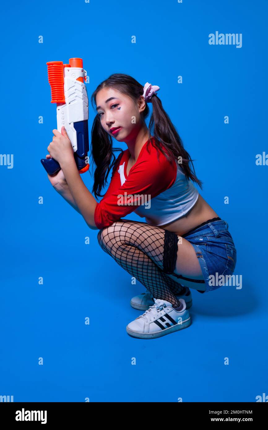 Mujer joven en Harlequin Traje cuclillas con pistola de agua en manos sobre fondo azul sólido Foto de stock