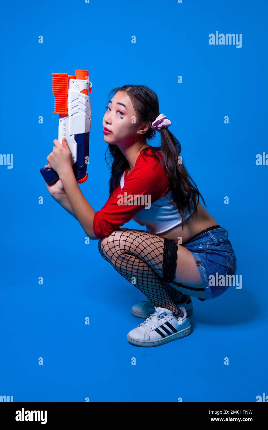 Mujer joven en Harlequin Traje cuclillas con pistola de agua en manos sobre fondo azul sólido Foto de stock