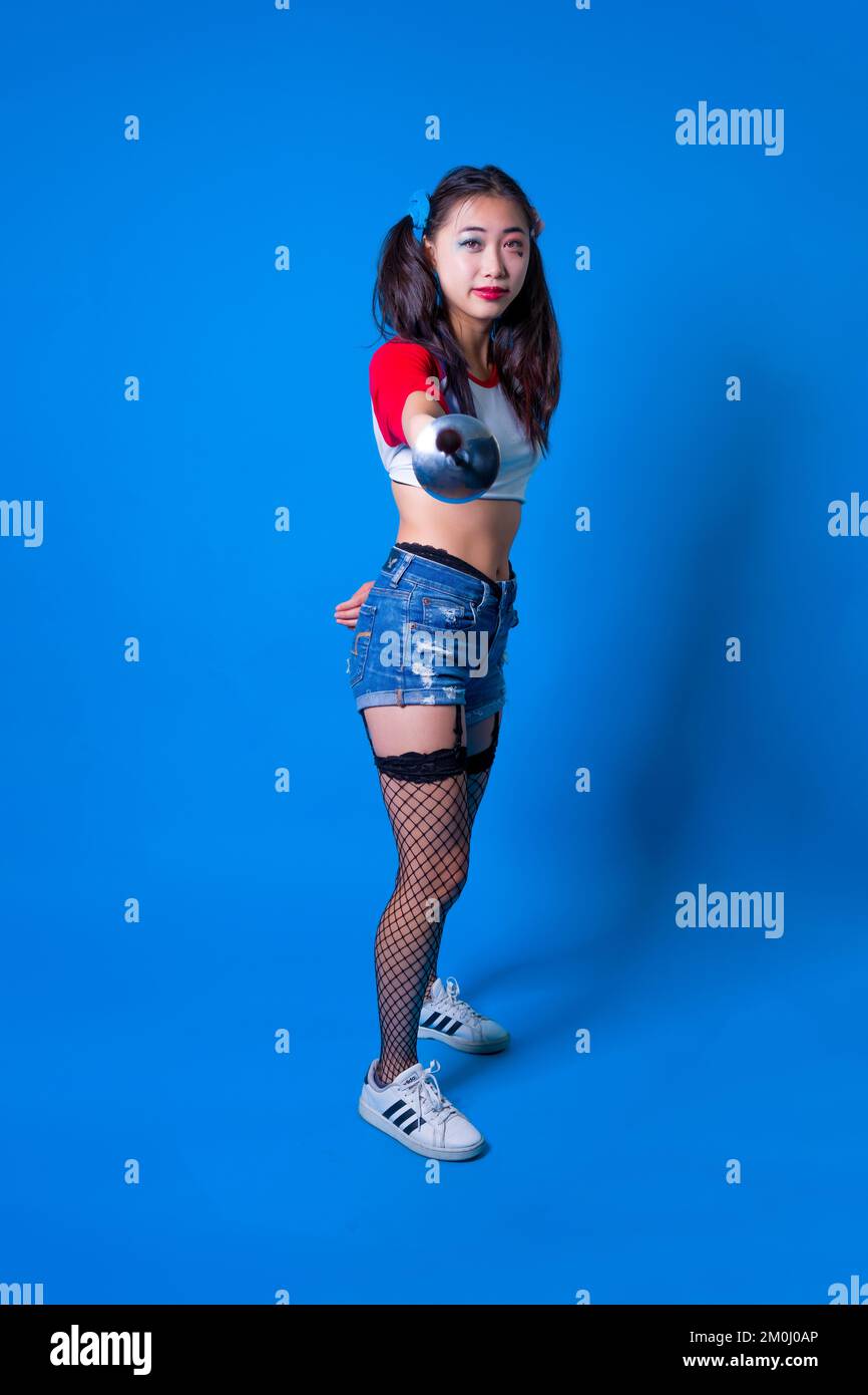 Mujer joven en traje Harlequin sosteniendo una película de esgrima contra un fondo azul sólido Foto de stock