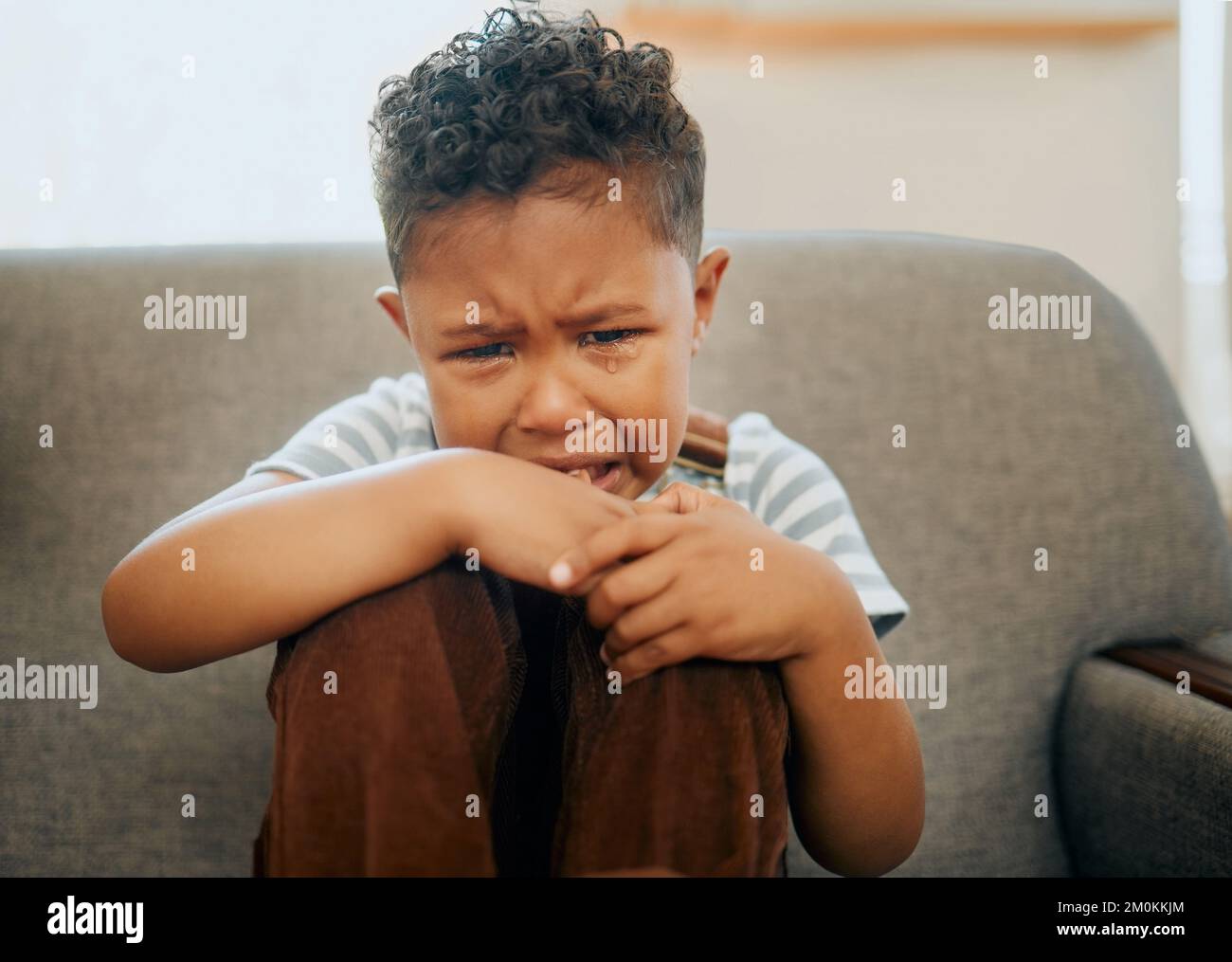 Un niño de carreras mixtas llorando en el sofá de la casa. Niño afroamericano que sufre de PTSD después de haber sido abusado y descuidado. Afligiendo poco Foto de stock