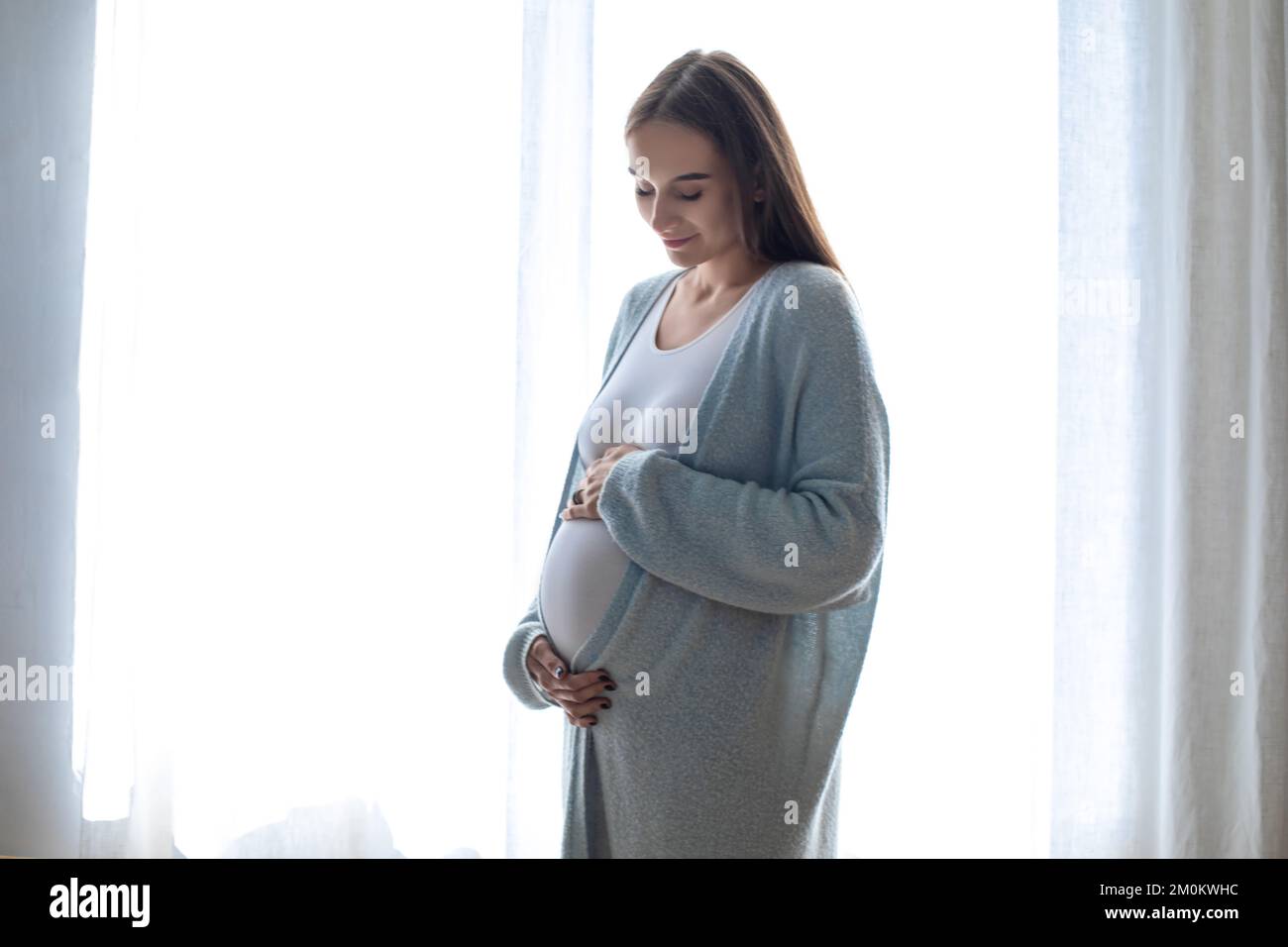 Mujer embarazada de pelo largo de pie cerca de la ventana Foto de stock