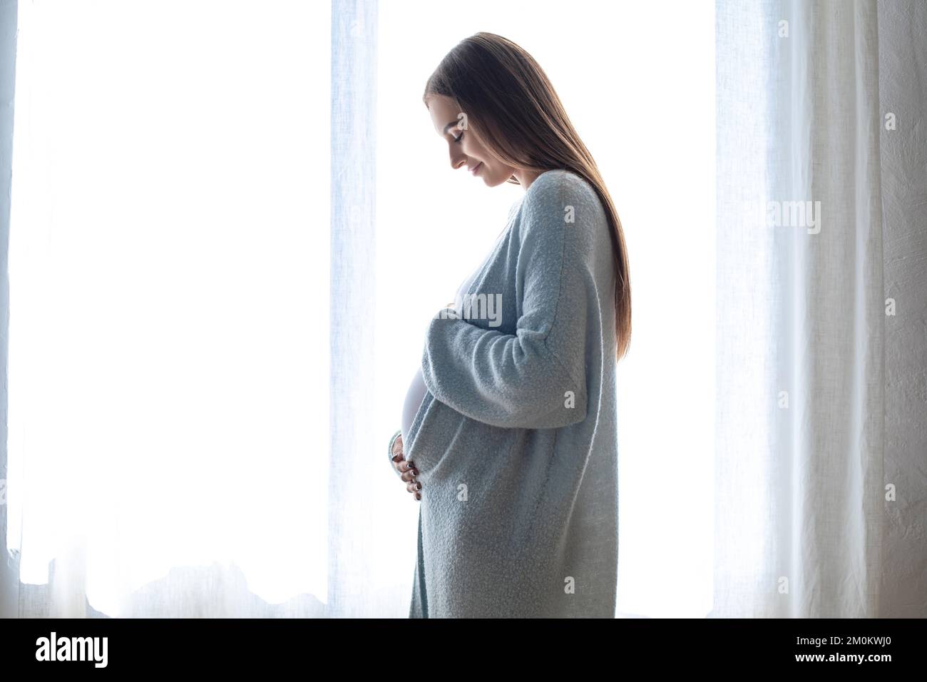 Mujer embarazada de pelo largo de pie cerca de la ventana Foto de stock