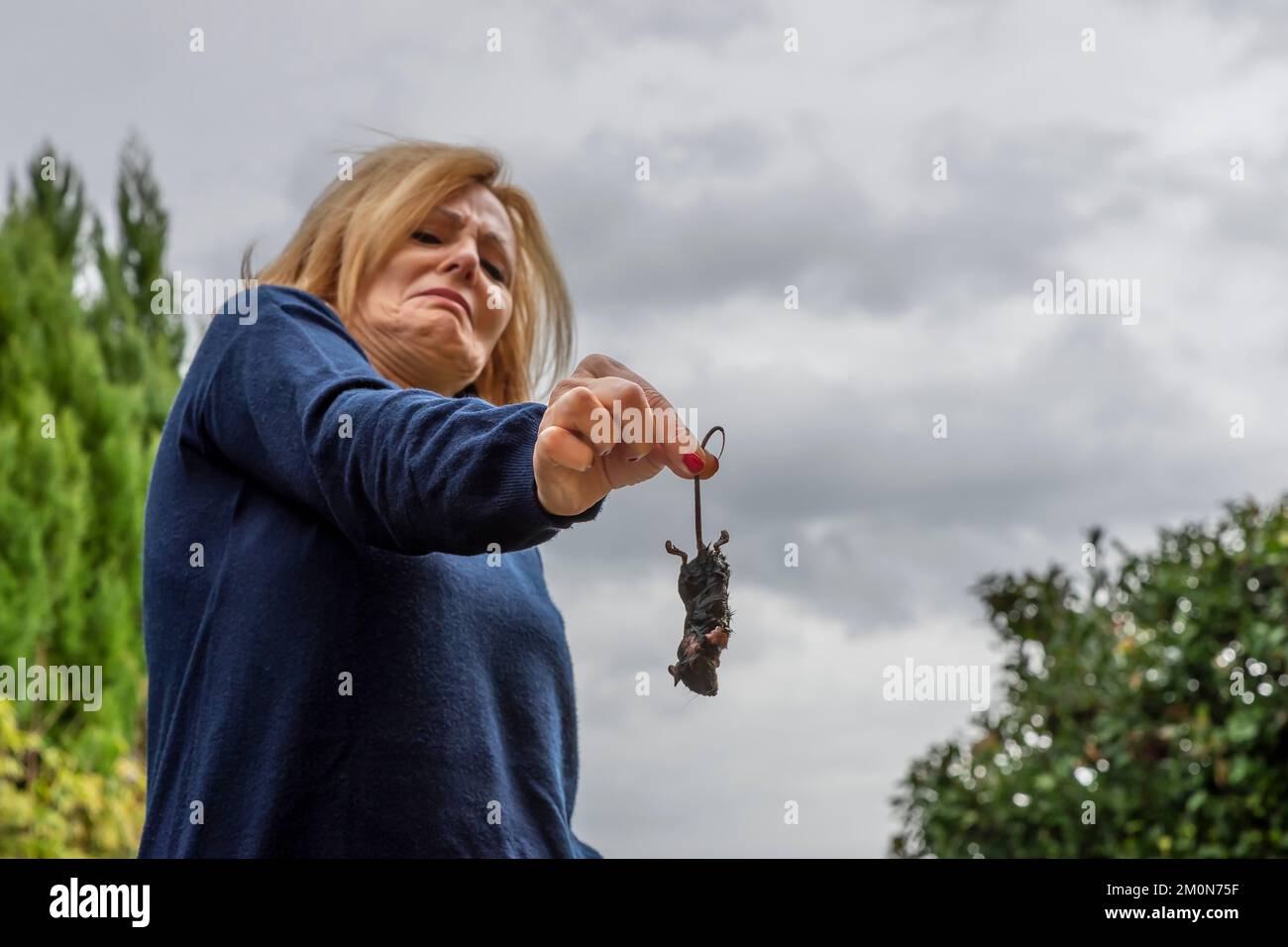 Una mujer rubia de mediana edad sostiene una rata mojada por la cola con una expresión aterrorizada y disgustada Foto de stock