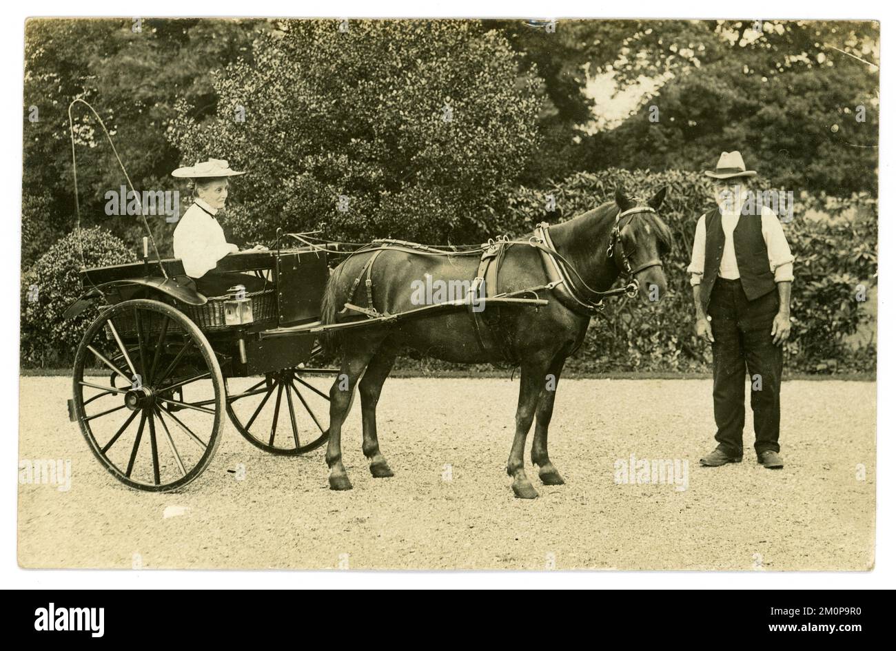 Original postal de principios de 1900 de una señora madura de clase alta que llevaba un sombrero grande de verano de moda sentado en un pony y atrapar con un sirviente, en la conducción de una casa grande, alrededor de 1910, Reino Unido Foto de stock