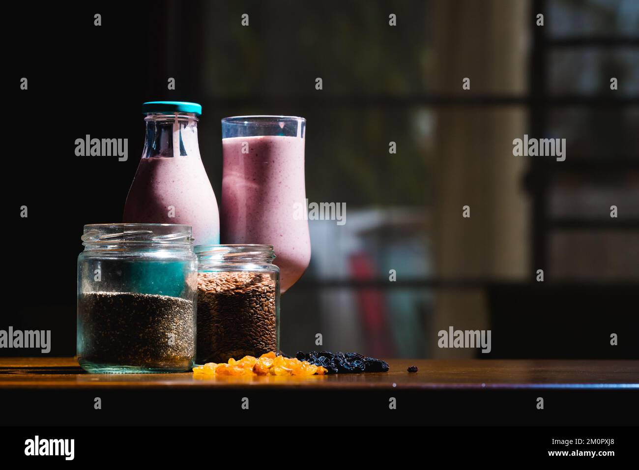 Una botella de vidrio con una taza de jugo, dos tarros de especias y frutas secas sobre una mesa de madera sobre un fondo borroso Foto de stock