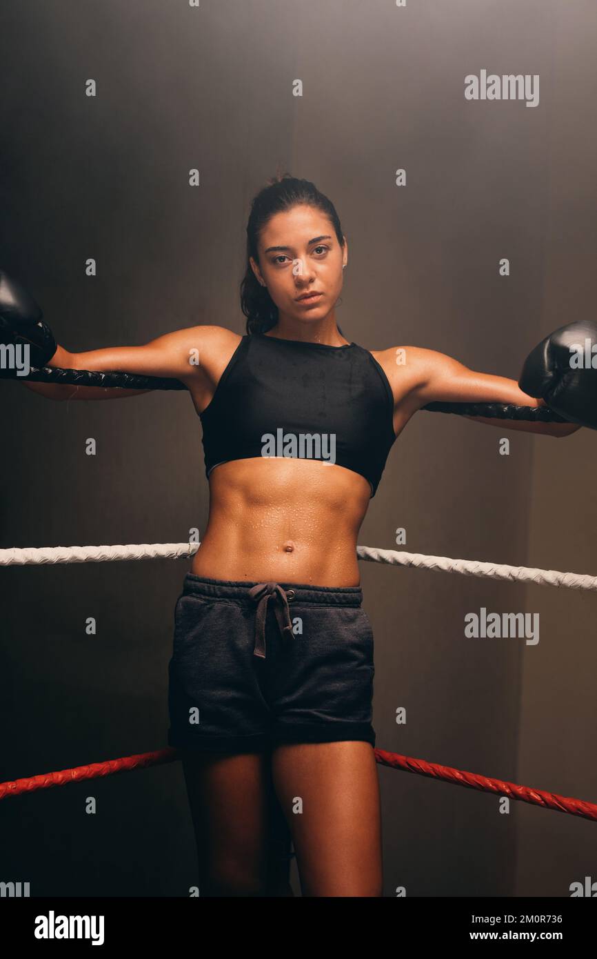 Joven boxer mirando la cámara mientras se apoya contra las cuerdas de un anillo de boxeo. Atleta mujer segura preparándose para un partido de boxeo. Foto de stock