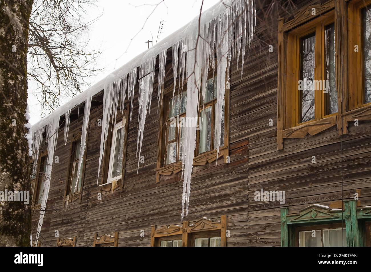 Largos y fundidos hielo cuelgan en el borde del techo, en invierno o en primavera. Muro de madera de una antigua casa de madera con ventanas. Grandes cascadas de hielo en suaves y hermosas filas. Día nublado de invierno, luz suave. Foto de stock