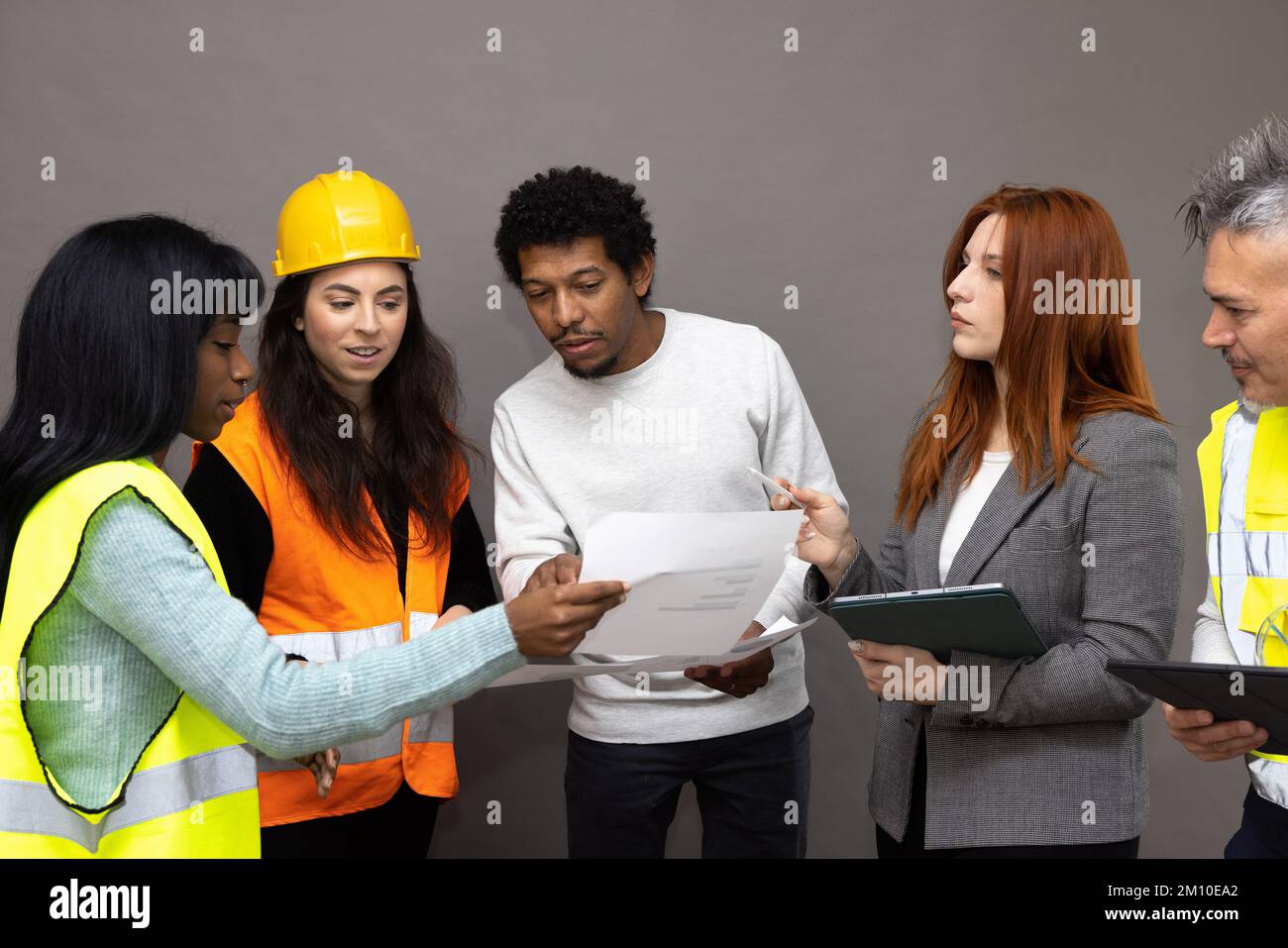 Grupo mixto de trabajadores del sitio de construcción observando y discutiendo el proyecto. Machos y hembras de diferentes etnias con chalecos de alta visibilidad Foto de stock