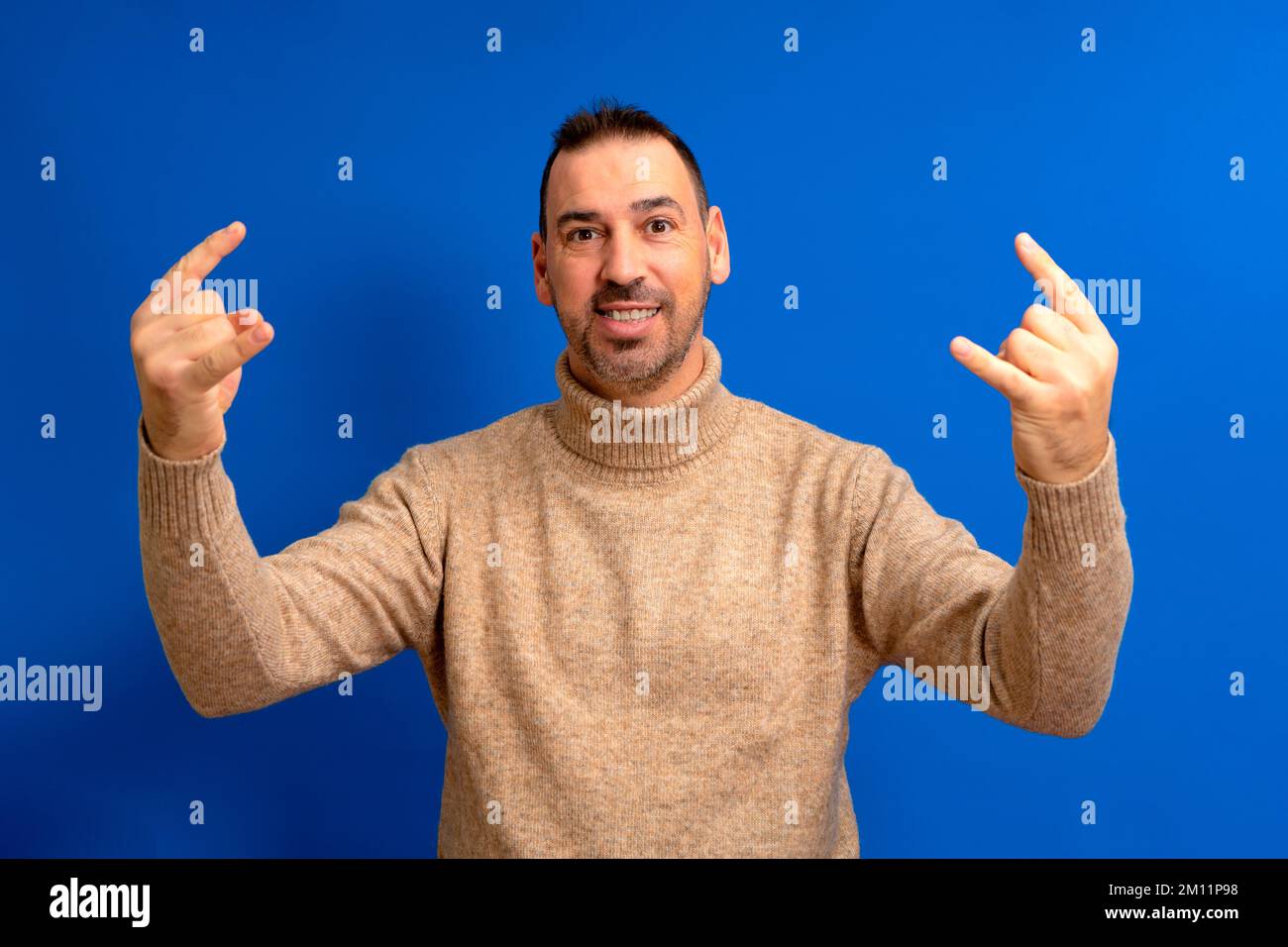 Un hombre latino barbudo que llevaba cuello de cuello alto marrón sobre un  fondo azul aislado gritando con una expresión loca haciendo un símbolo de  rock con las manos arriba. Estrella de