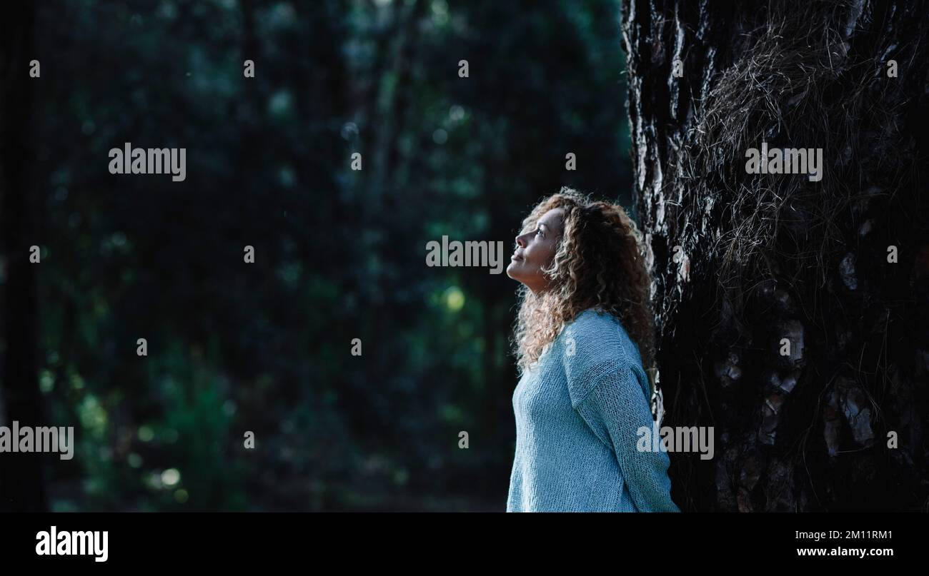 Imagen oscura de una mujer de pie contra un tronco de árbol en la naturaleza exterior actividad de ocio con bosques en el fondo. Una mujer disfruta mirando al bosque y relajándose. Entorno en el parque. Espacio de copia Foto de stock