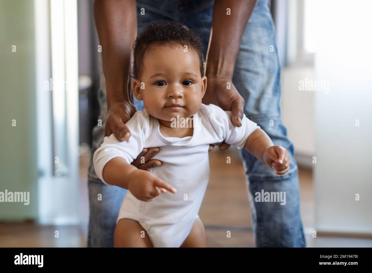Retrato del adorable pequeño bebé negro caminando con la ayuda del padre en la habitación Foto de stock