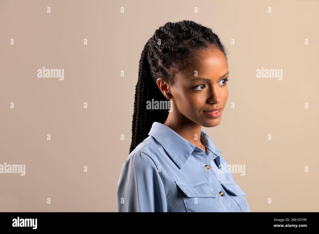 Joven modelo femenino negro con trenzas afro y camisa azul sobre fondo beige mirando a distancia Foto de stock