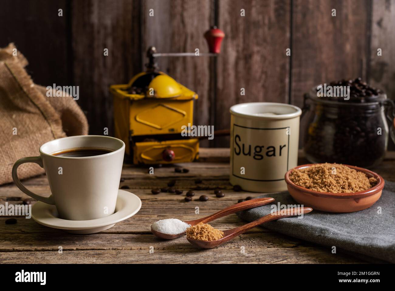 Taza de café recién hecho junto a unas cucharas de madera con azúcar blanco  y otra con azúcar morena y sus jarras detrás, en el fondo un viejo café  Fotografía de stock -