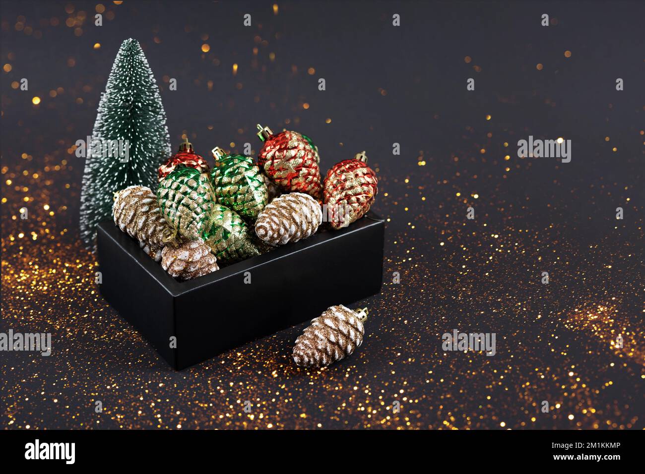 Decoraciones de Navidad en forma de conos de pino rojo, amarillo y verde en una caja negra con un pequeño árbol decorativo de Navidad sobre un fondo negro con Foto de stock
