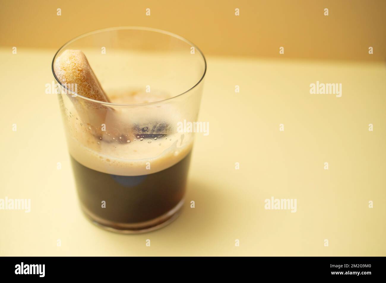 Galleta ladyfinger esponjosa sumergida en café espresso, en un vaso transparente, sobre fondo de color crema. Enfoque suave de primer plano. Foto de stock