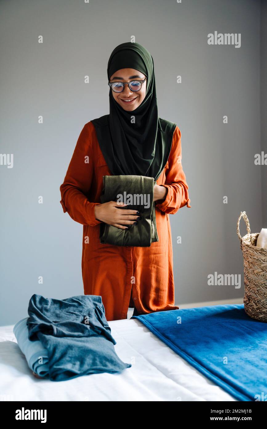 Joven hermosa mujer sonriente en hijab y gafas doblando la ropa en casa Foto de stock