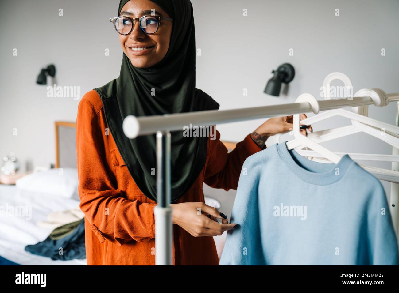 Joven hermosa mujer sonriente en hijab y gafas colgando ropa en la barandilla en casa Foto de stock