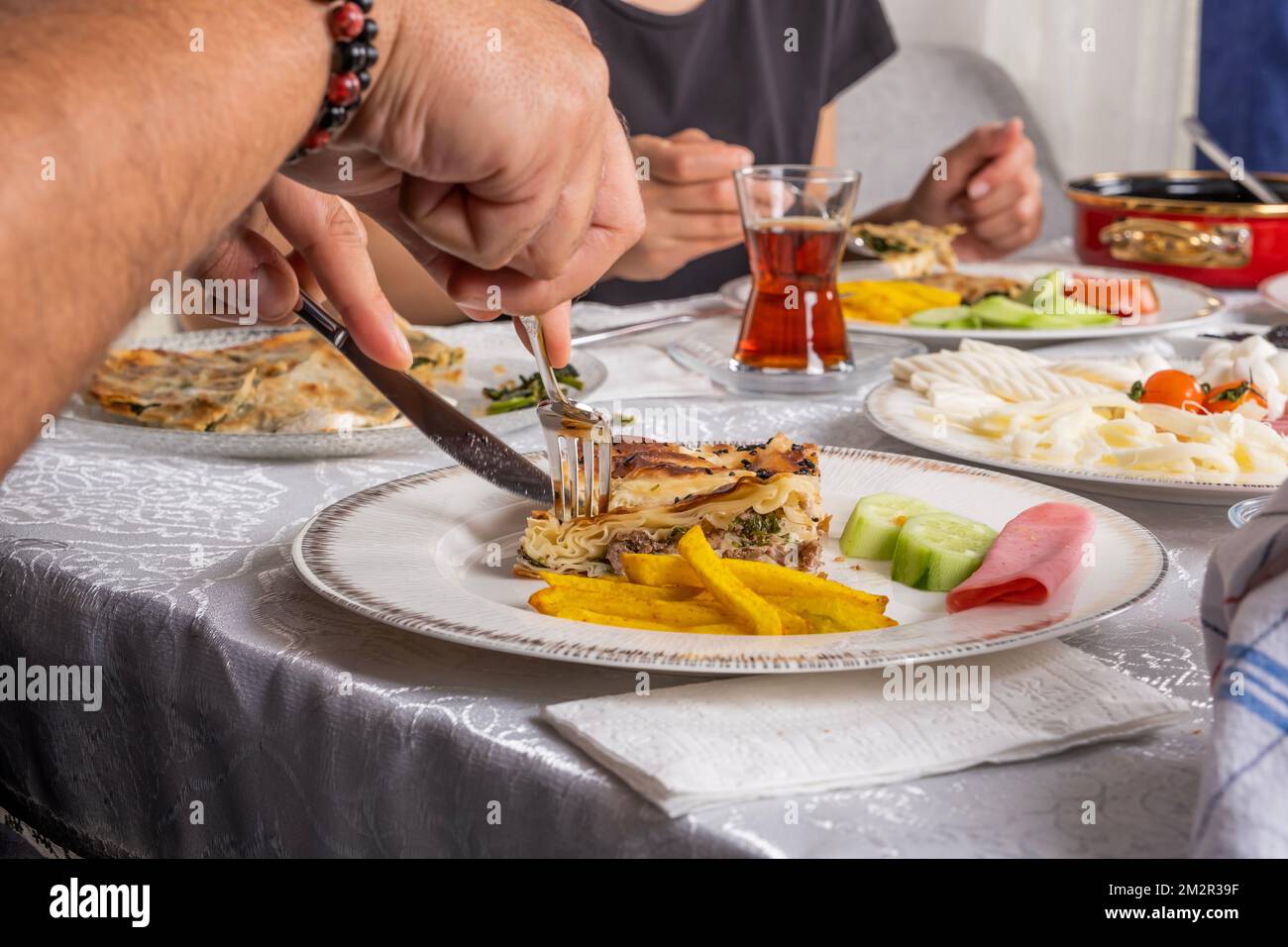 Cerrar la imagen de las manos masculinas cortando la torta turca llamada su borek con el cuchillo. Mesa de desayuno tradicional. Té en vaso. Rutina de domingo por la mañana. Foto de stock