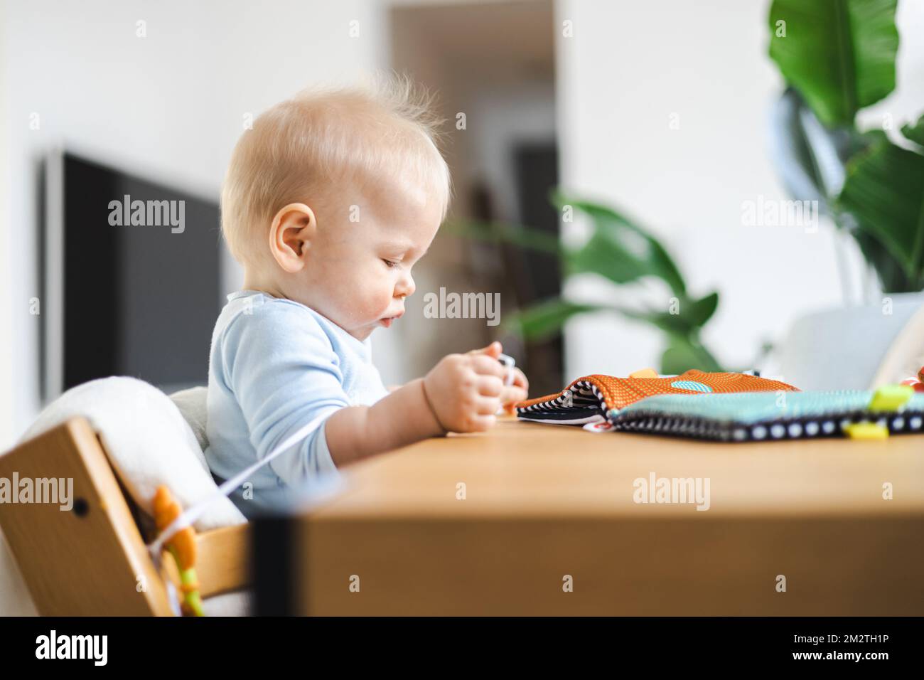 Bebé feliz sentado en la mesa del comedor y jugando con su juguete en la silla alta de madera de diseño escandinavo tradicional en una casa moderna y luminosa. Lindo bebé jugando con juguetes. Foto de stock
