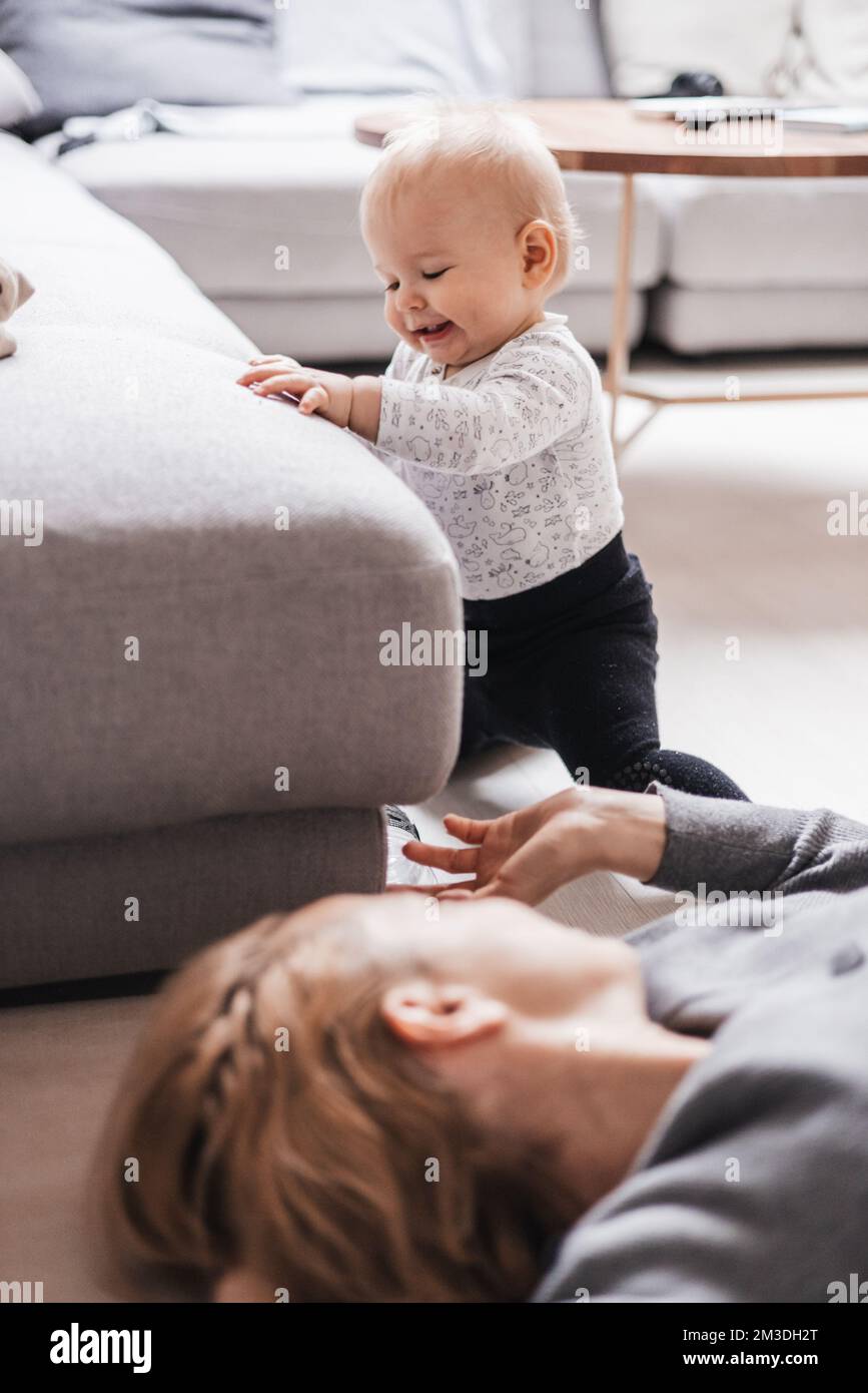 Felices momentos en familia. Madre tumbada cómodamente en la colchoneta de los niños jugando con su bebé mirando y superando sus primeros pasos. Emociones humanas positivas, sentimientos, alegría Foto de stock