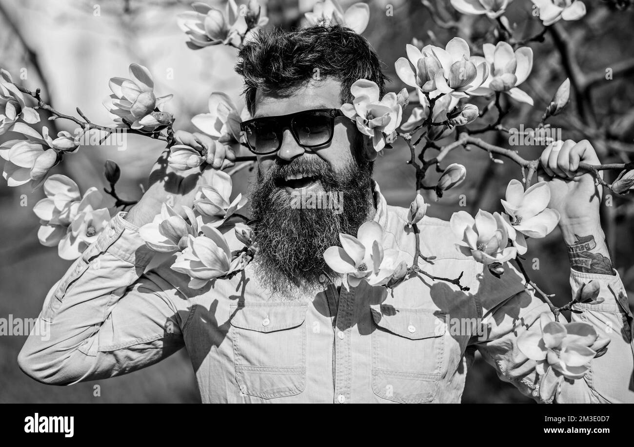 Hipster en gafas de sol de moda alegremente posando con ramas de magnolia  en el fondo. Guy en gafas de sol modernas con estilo. Concepto de moda.  Hombre con Fotografía de stock -
