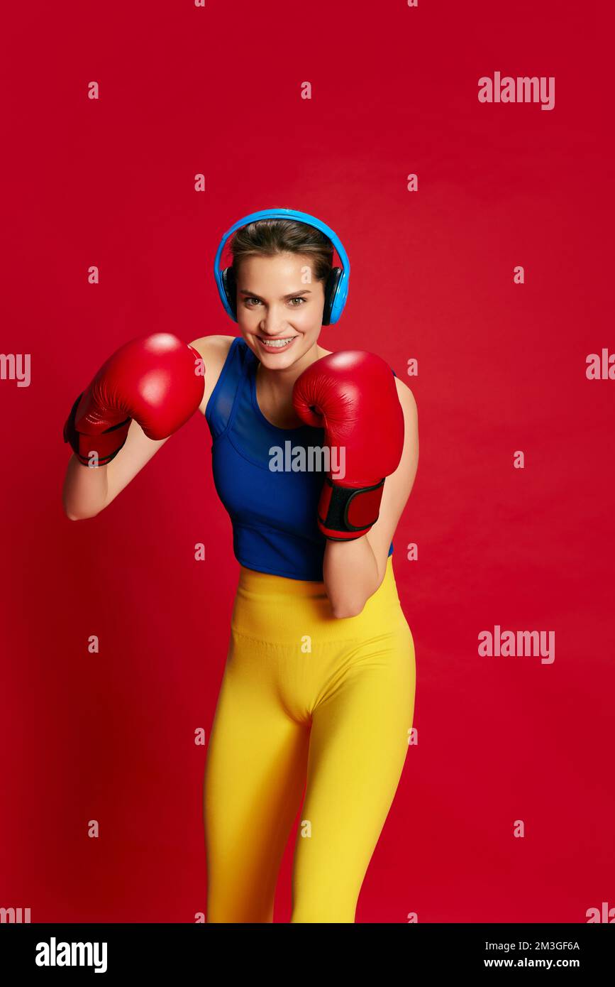 Retrato de joven hermosa chica en ropa deportiva, auriculares posando  guantes de boxeo inn sobre fondo rojo. Fuerza Fotografía de stock - Alamy
