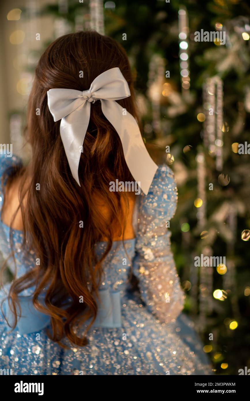 La chica decora el árbol de Navidad, sosteniendo bolas de Navidad en su  mano. Ella usa un vestido azul con un arco en su cabello. Concepto de  vacaciones Fotografía de stock -