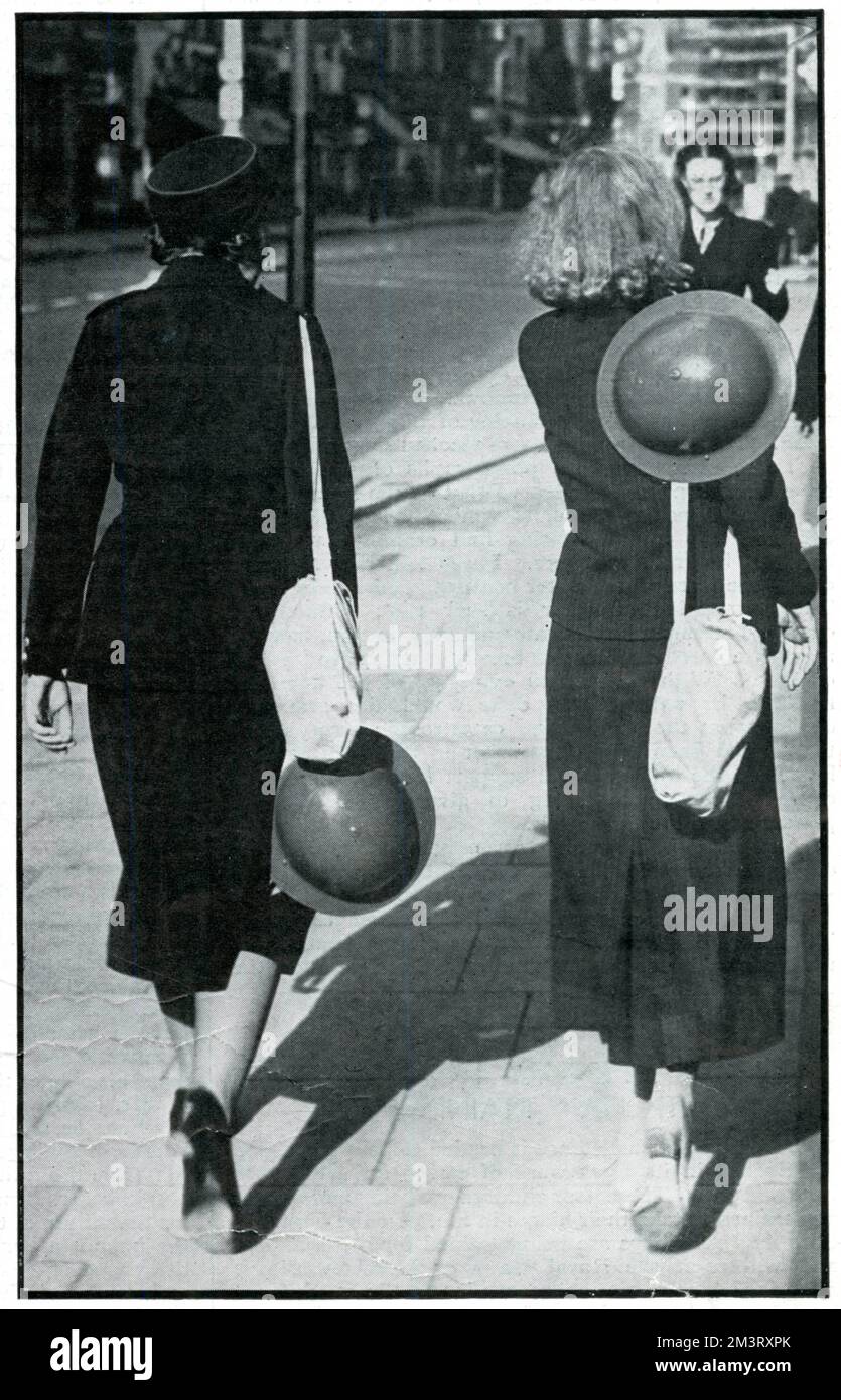 Dos chicas, descritas en uniformes elegantes, fotografiadas por detrás mientras caminaban por una calle londinense sosteniendo cascos de acero. La Esfera comenta el cambio de moda en tiempos de guerra en las semanas previas al estallido de la guerra. 1939 Foto de stock