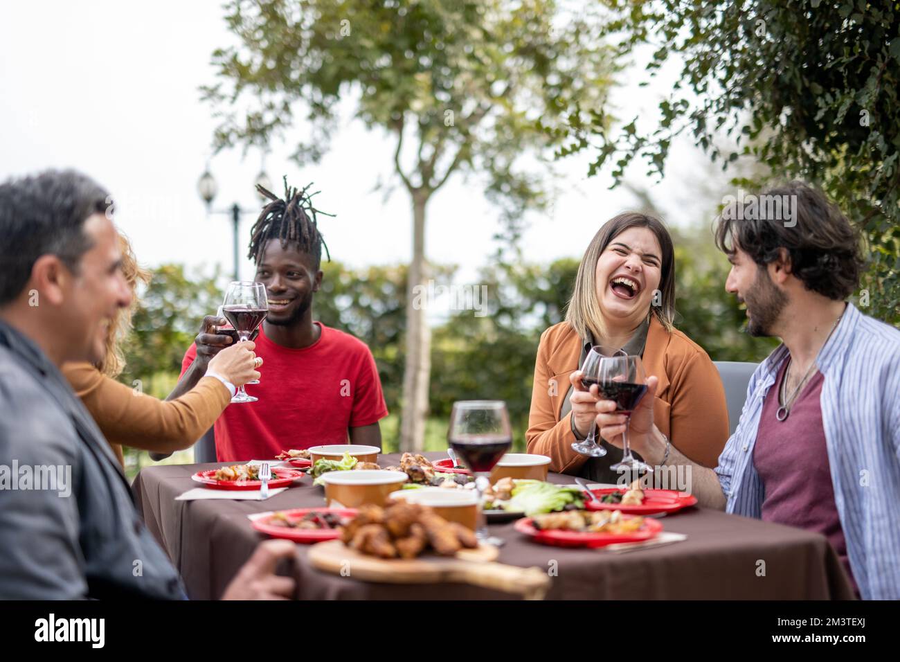 Gente feliz que tiene buenos momentos mientras que come la comida y bebe el vino en el jardín, la diversidad y el concepto de rango de edad mezclado Foto de stock