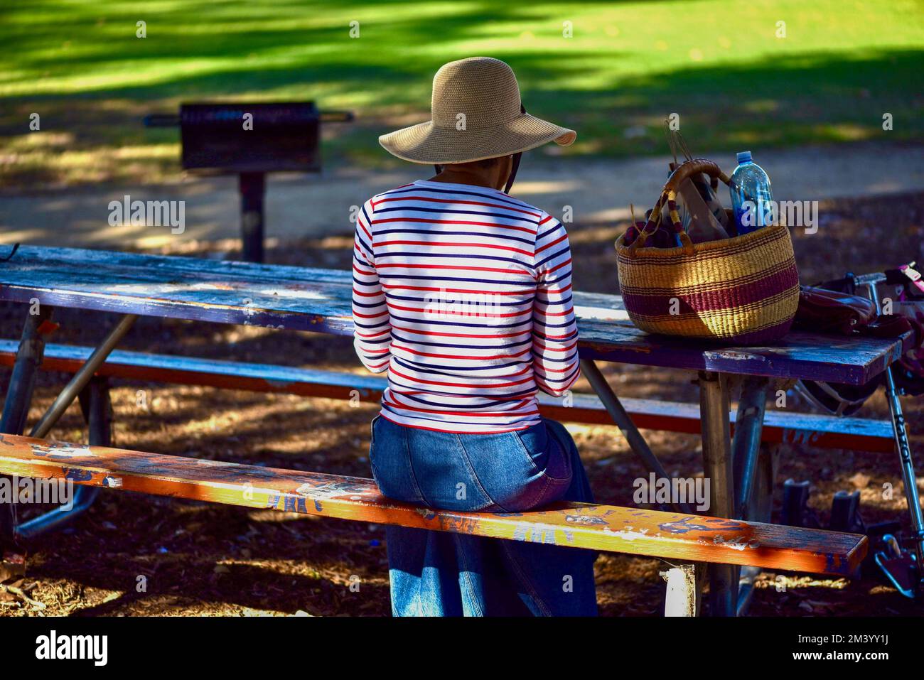 la mujer sentada en un banco del parque que lleva un sombrero floppy de fibra natural tiene un almuerzo tranquilo y silencioso en un entorno similar al parque Foto de stock
