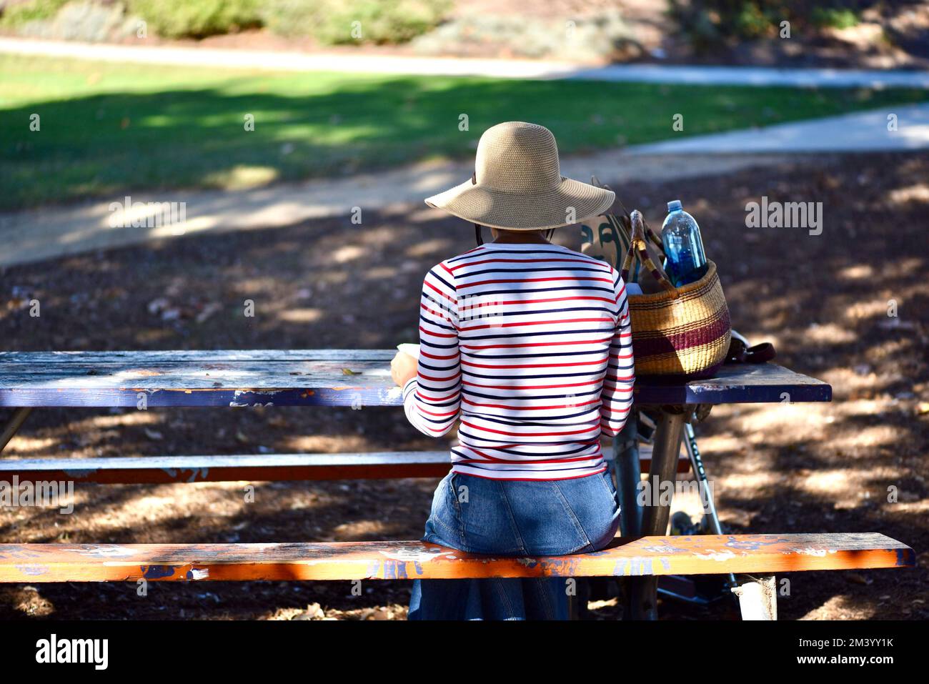 la mujer sentada en un banco del parque que lleva un sombrero floppy de fibra natural tiene un almuerzo tranquilo y silencioso en un entorno similar al parque Foto de stock