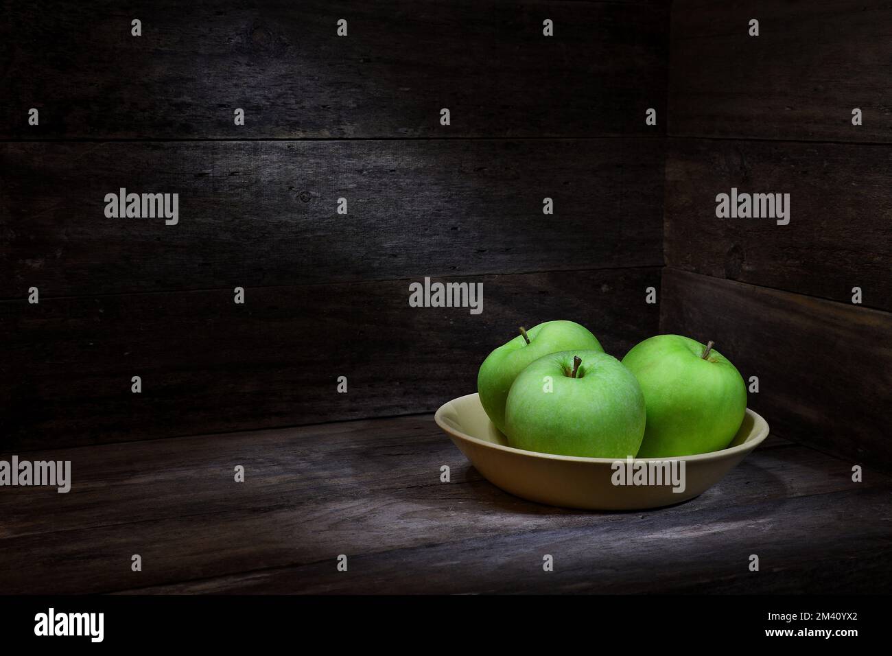 Una escena rústica de tres manzanas verdes Granny Smith en un tazón amarillo en una caja de madera que rodea con iluminación de humor oscuro; capturado en un estudio Foto de stock