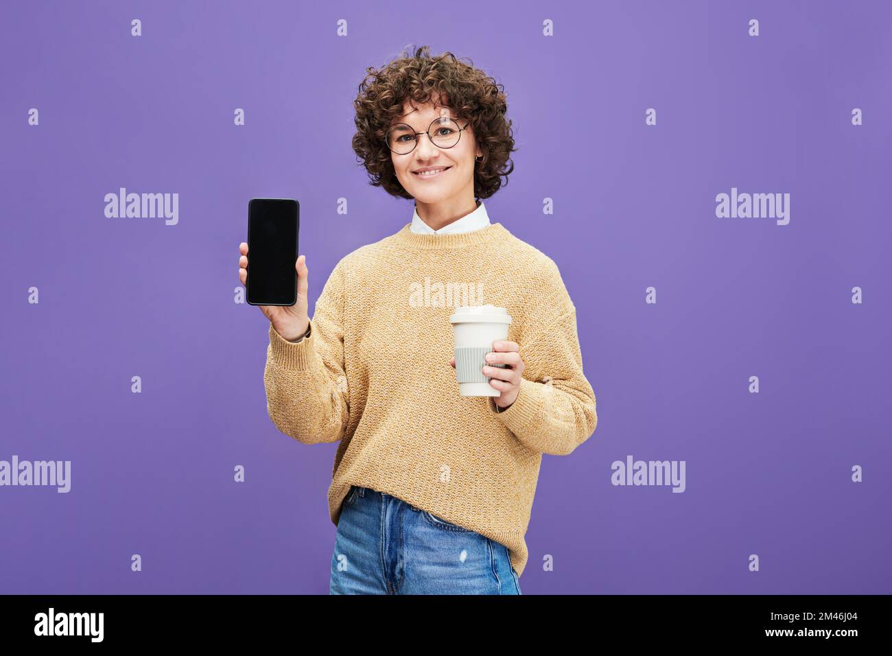 Mujer morena joven feliz con la taza de café que muestra el smartphone con la pantalla en blanco donde usted puede poner el texto del anuncio Foto de stock